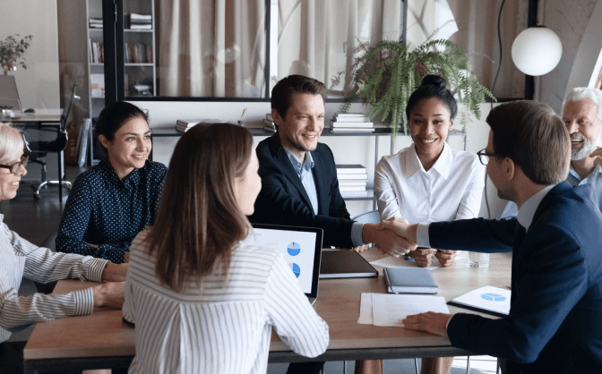 Transferencia Temporal Valores Foto de un grupo de empresarios, entre hombres y mujeres reunidos en la mesa de una oficina, dos de los hombres se dan la mano para hablar sobre los beneficios de la Transferencia Temporal Valores.