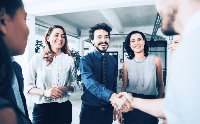 Crecimiento de capital Foto de un grupo de personas reunidas sonriendo en la oficina, dos hombres se dan la mano y hablan sobre inversiones para obtener crecimiento de capital en Valores Bancolombia.