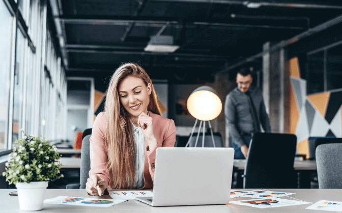 Inversión inmobiliaria Foto de una mujer joven con cabello largo, sonriendo en su escritorio donde está su computador portátil y mirando los documentos con los resultados de la inversión inmobiliaria con Valores Bancolombia.
