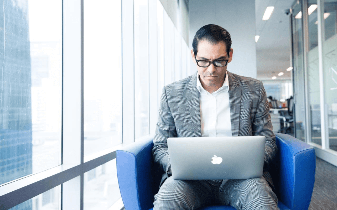 Alternativas de inversión Foto de un hombre con gafas y traje sentado en un mueble con un computador portátil en sus piernas leyendo sobre las alternativas de inversión en Valores Bancolombia.